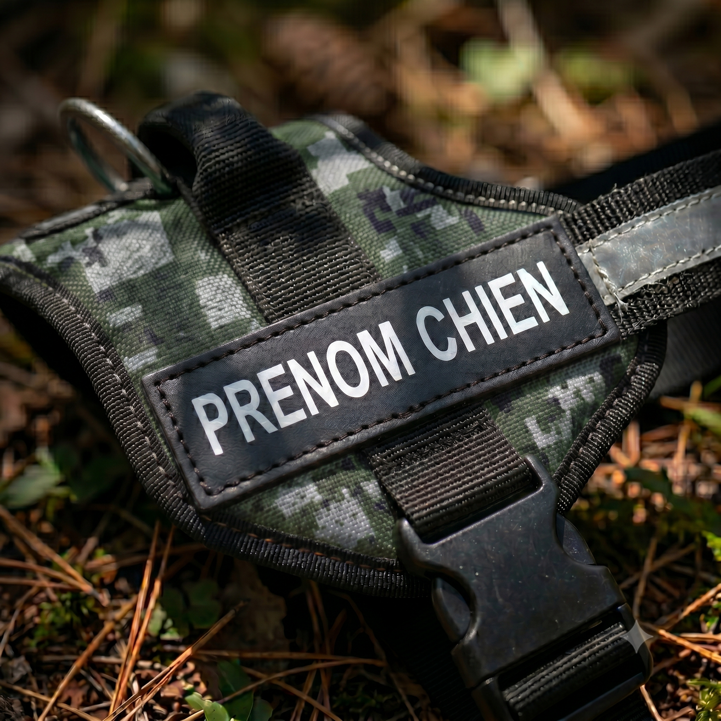 Gros plan sur le patch prénom réfléchissant d'un harnais chien personnalisé motif camouflage posé en extérieur, avec boucle de sécurité et anneau de laisse